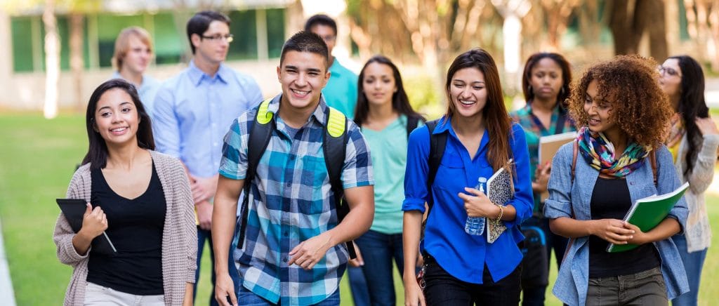 Teenagers or young adults walking outdoors on school campus. Hispanic, Caucasian, and African American students are smiling while walking together to class. They are wearing trendy casual clothing and backpacks, and are carrying school books.