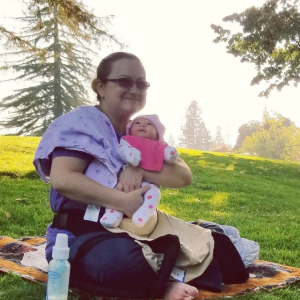 A woman sits on a picnic blanket with her newborn baby. It's a sunny day at the park.