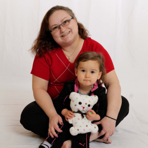 A woman poses with a toddler holding a teddy bear against a white backdrop. 