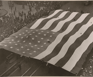 A black and white photo shows a large group of people carrying a massive American flag in a parade.