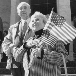 A black and white photo depicts an older couple brandishing American flags in celebration of their newfound citizenship.