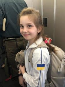 A young girl with a Ukrainian flag on her shirt holds a teddy bear as she boards a plane.