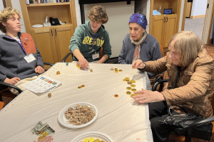 A senior and a group of teen volunteers sit around a table playing dreidel and smiling