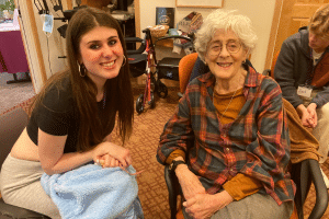 A female teen volunteer sitting next to a senior citizen, both smiling broadly